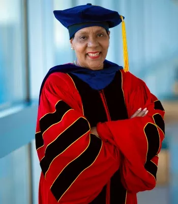 Diane Howard, PhD, wearing a graduation cap and gown, standing indoors in front of a row of windows
